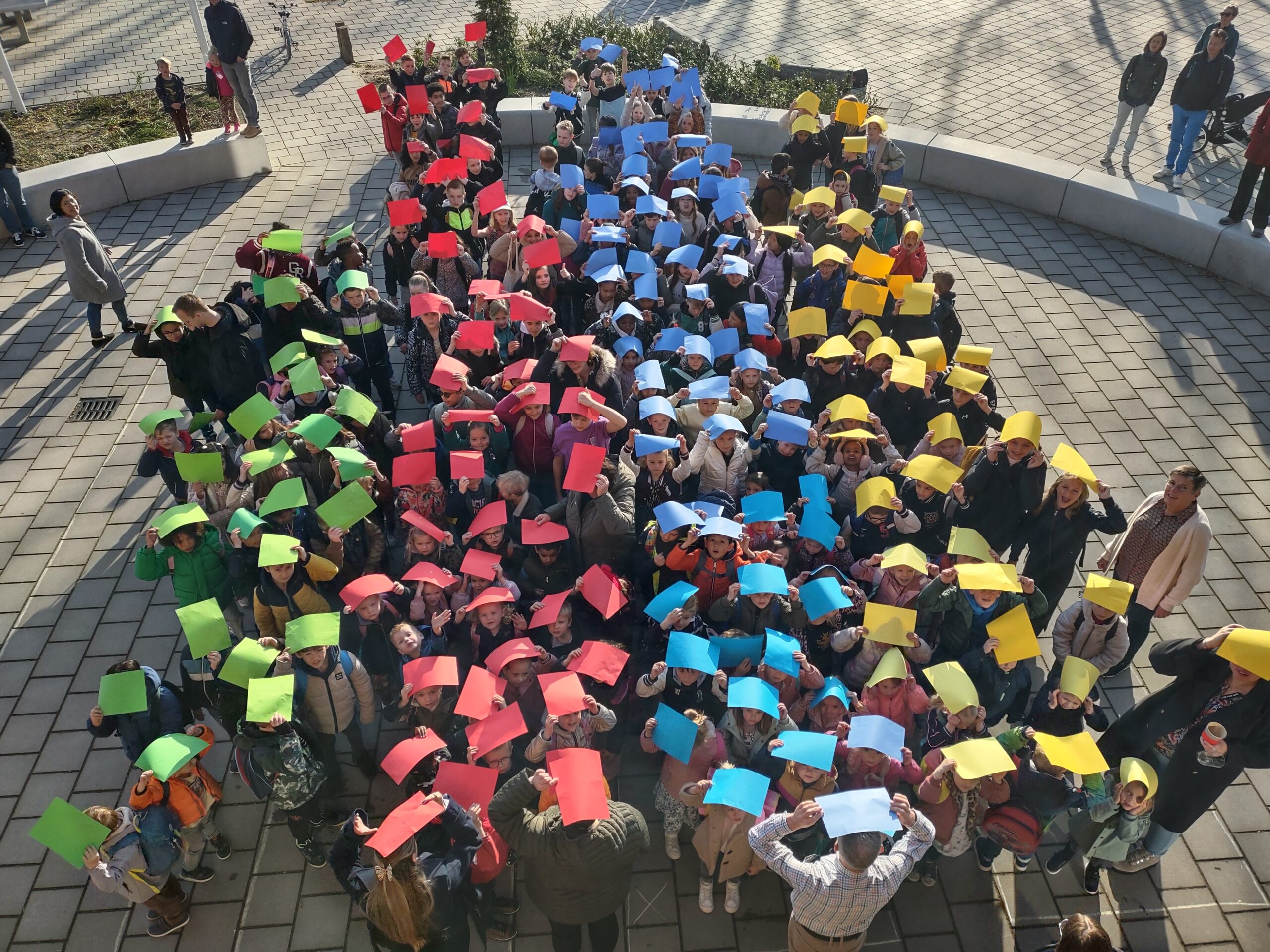 Leerlingen met een gekleurd papier boven hun hoofd vormen een grafiek op het plein